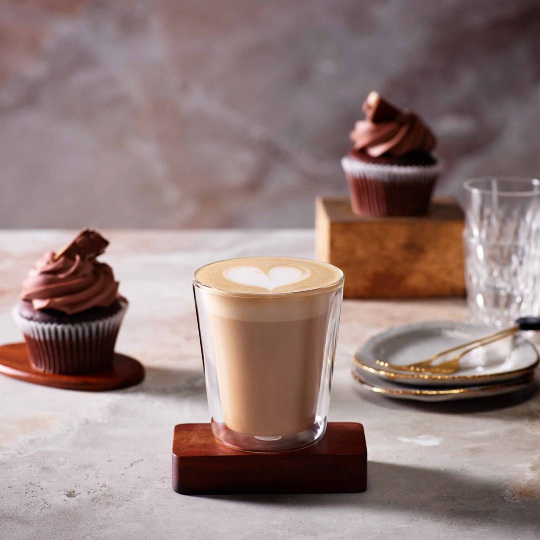 A latte glass with a heart design on top, surrounded by cupcakes and plates