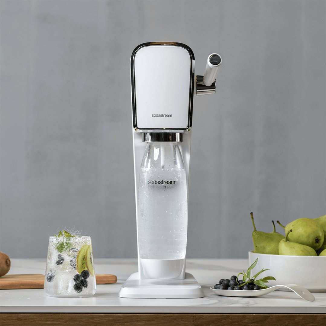 A white SodaStream Art Sparkling Water Maker on a counter with fruit and a drink