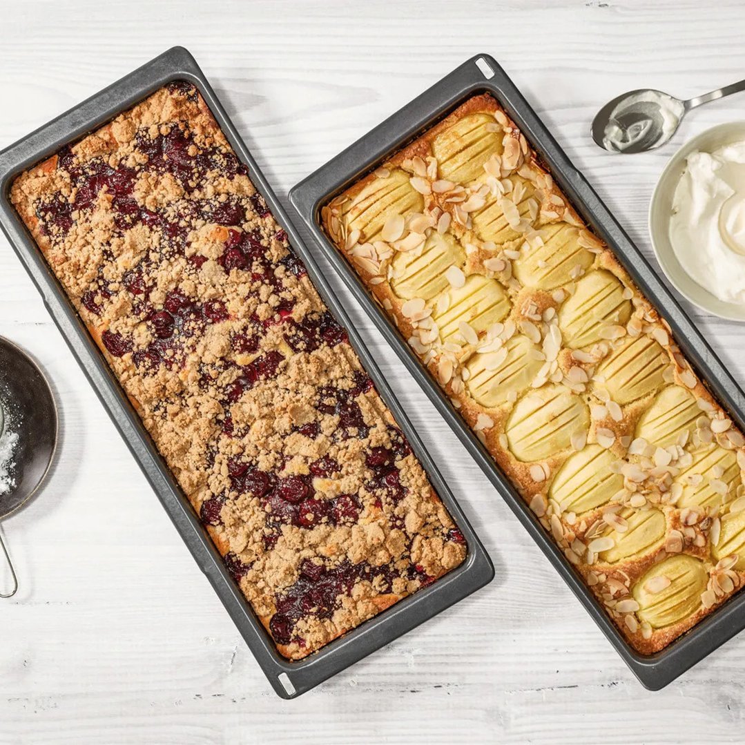Two slim universal pans with baked goods on a white wooden surface