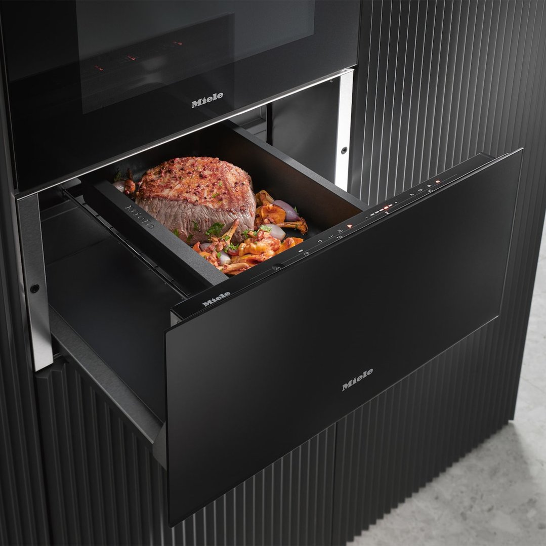 Open warming drawer with a cooked roast and vegetables inside, built into a black cabinet